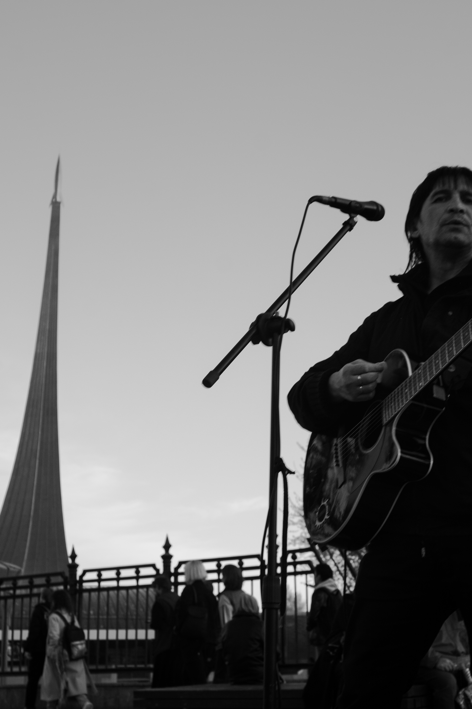 Black and white photo of a musician playing guitar outdoors near a tall monument.