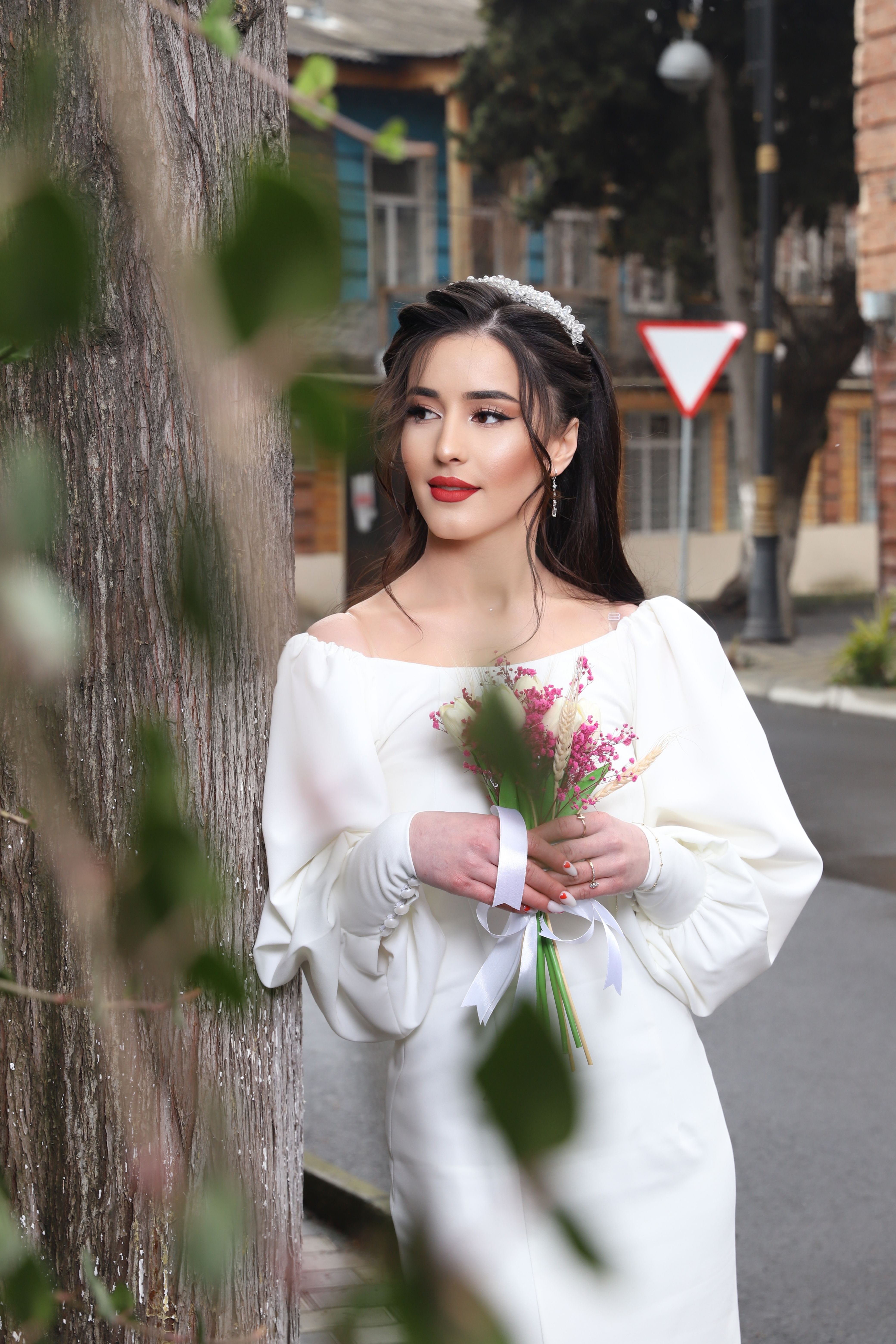 A stylish woman in a white dress poses outdoors, holding a bouquet near a tree.