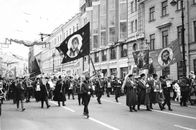 Black and white image of people marching in a city with flags and banners.
