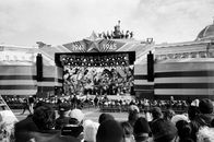 Black and white image of a crowd watching a historical stage performance outdoors.