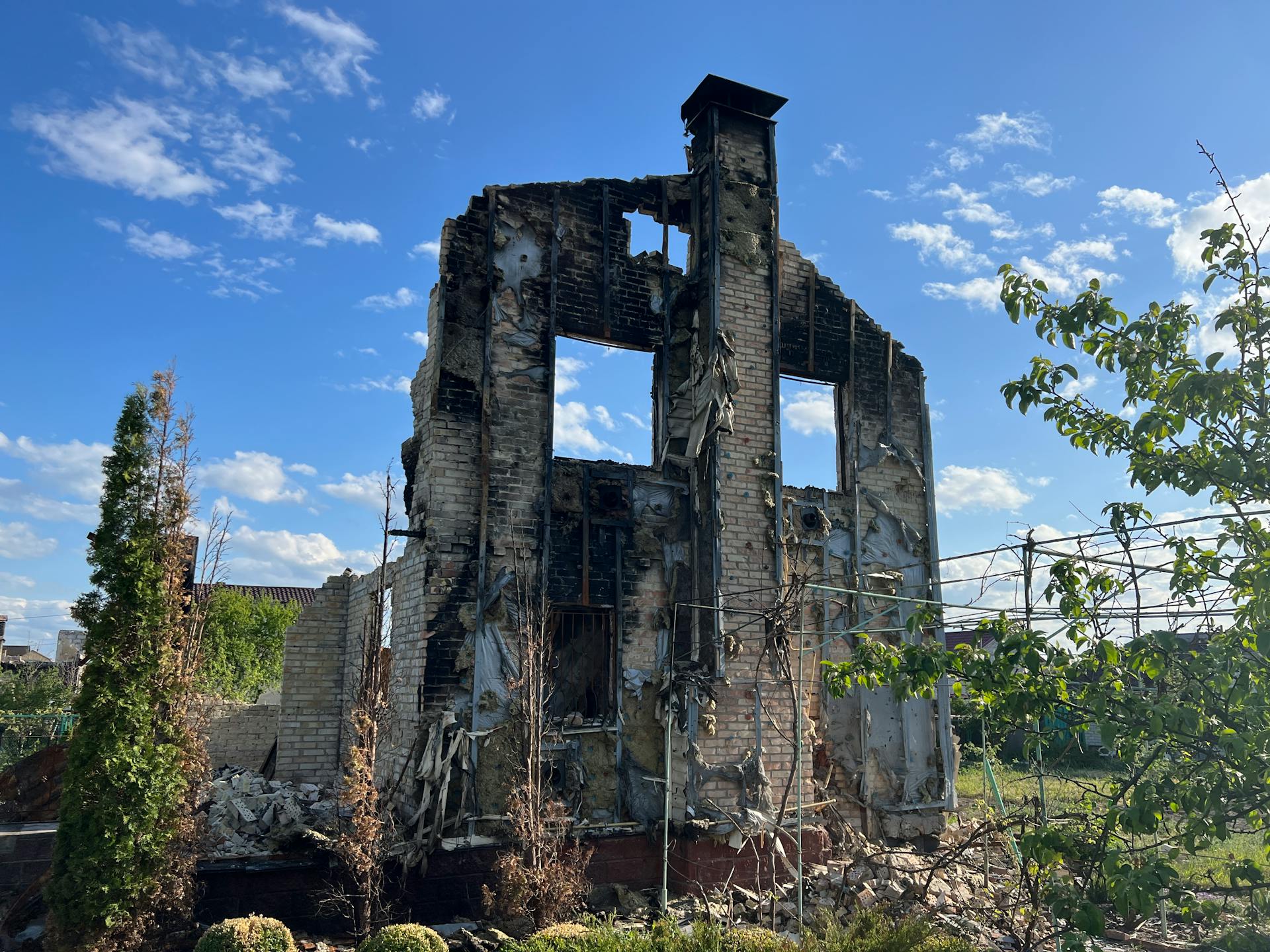 A desolated, burned-down house stands beneath a vibrant blue sky, symbolizing destruction and resilience.