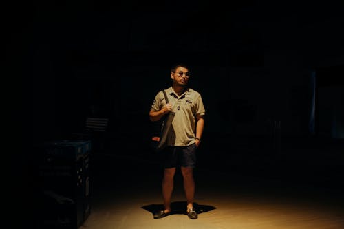 Free Man in Brown Polo Shirt and Black Shorts Standing in a Dark Room Carrying a Bag Stock Photo