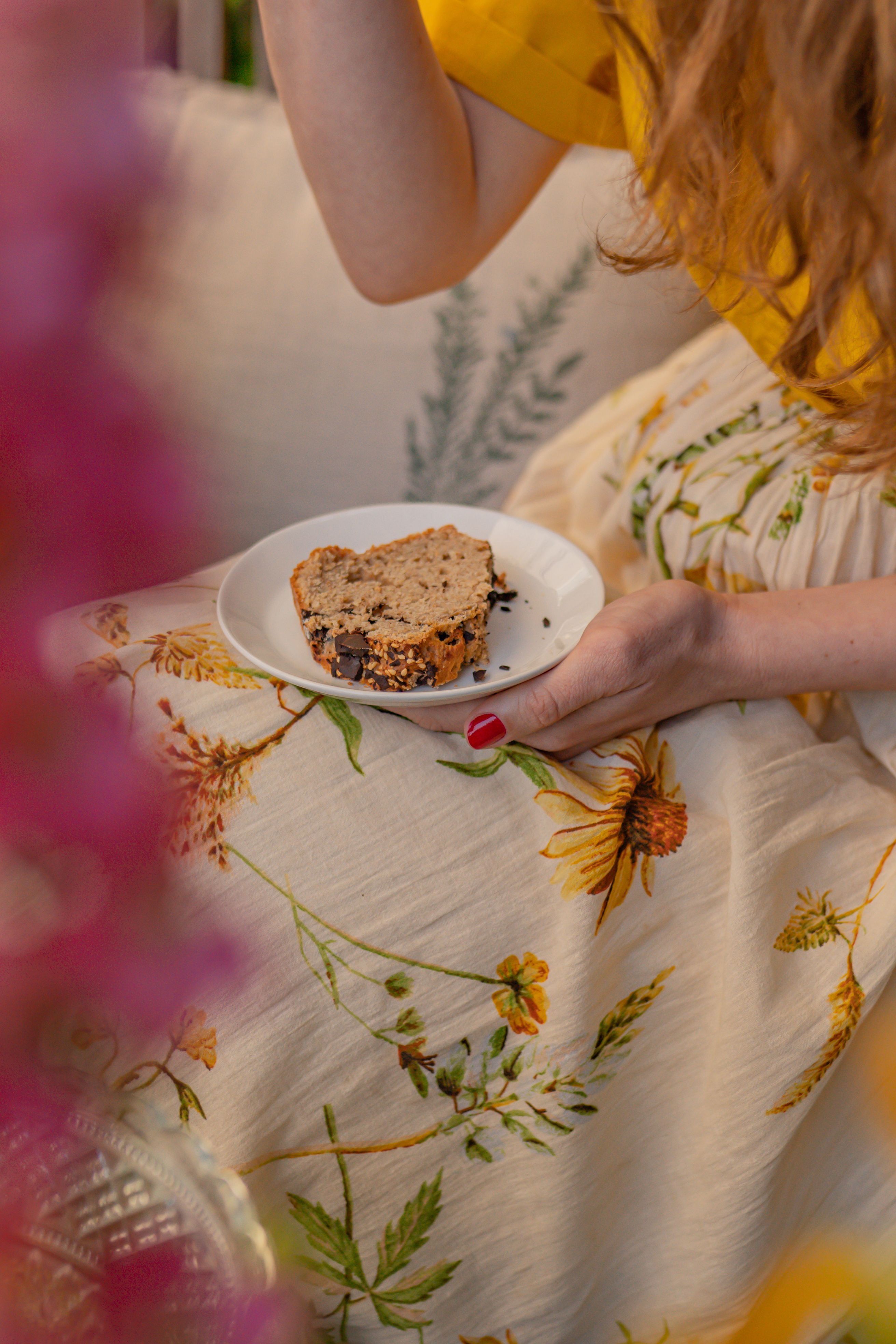 Close-up of woman holding a piece of cake on a floral dress, enjoying a sunny outdoor setting.