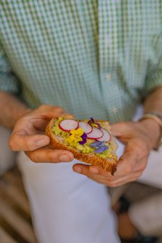 Fresh avocado toast topped with radishes and edible flowers held by a person in a green shirt.