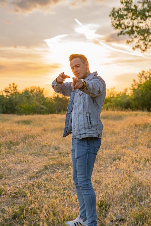 Man in Denim Jacket Standing on Grass Field during Sunset