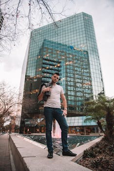 A confident man poses by a plaza fountain in front of a modern skyscraper, exuding urban charm.