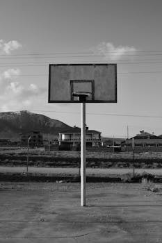 Grayscale image of an outdoor basketball hoop against a mountainous backdrop.