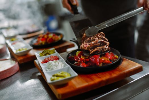 Close-up of a chef preparing grilled steak with vegetables and sauces on a wooden board in a restaurant kitchen.