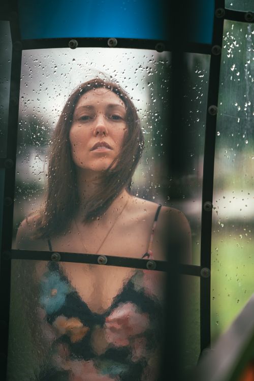 Free A woman gazes through a rain-speckled window, creating a reflective and moody atmosphere. Stock Photo