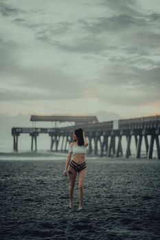 Woman in swimwear standing on a beach with a pier in the background.