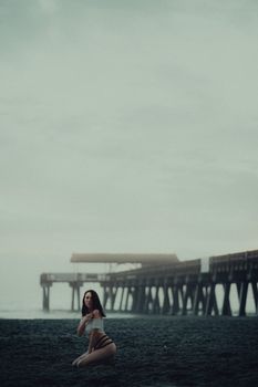 A woman in swimwear sits on a sandy beach beside a long ocean pier under a cloudy sky.