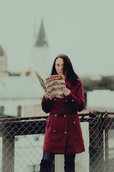 A woman in a stylish red coat stands outdoors reading a magazine, blending fashion with leisure.