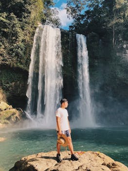 A man in shorts and a hat stands on rocks near a stunning waterfall surrounded by lush forest.