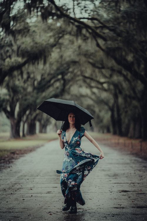 Free Woman in a blue floral dress walks with an umbrella down a tree-lined road on a rainy day. Stock Photo