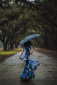 A woman in a floral dress dances with an umbrella along a scenic, tree-lined path during rain.