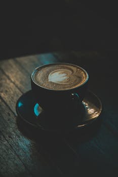 A cup of latte with intricate art on a wooden table in a dimly lit café setting.