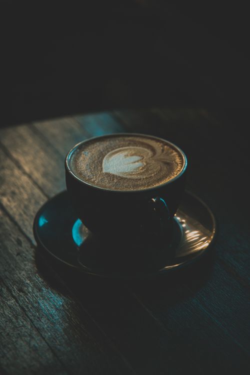 Free A cup of latte with intricate art on a wooden table in a dimly lit café setting. Stock Photo