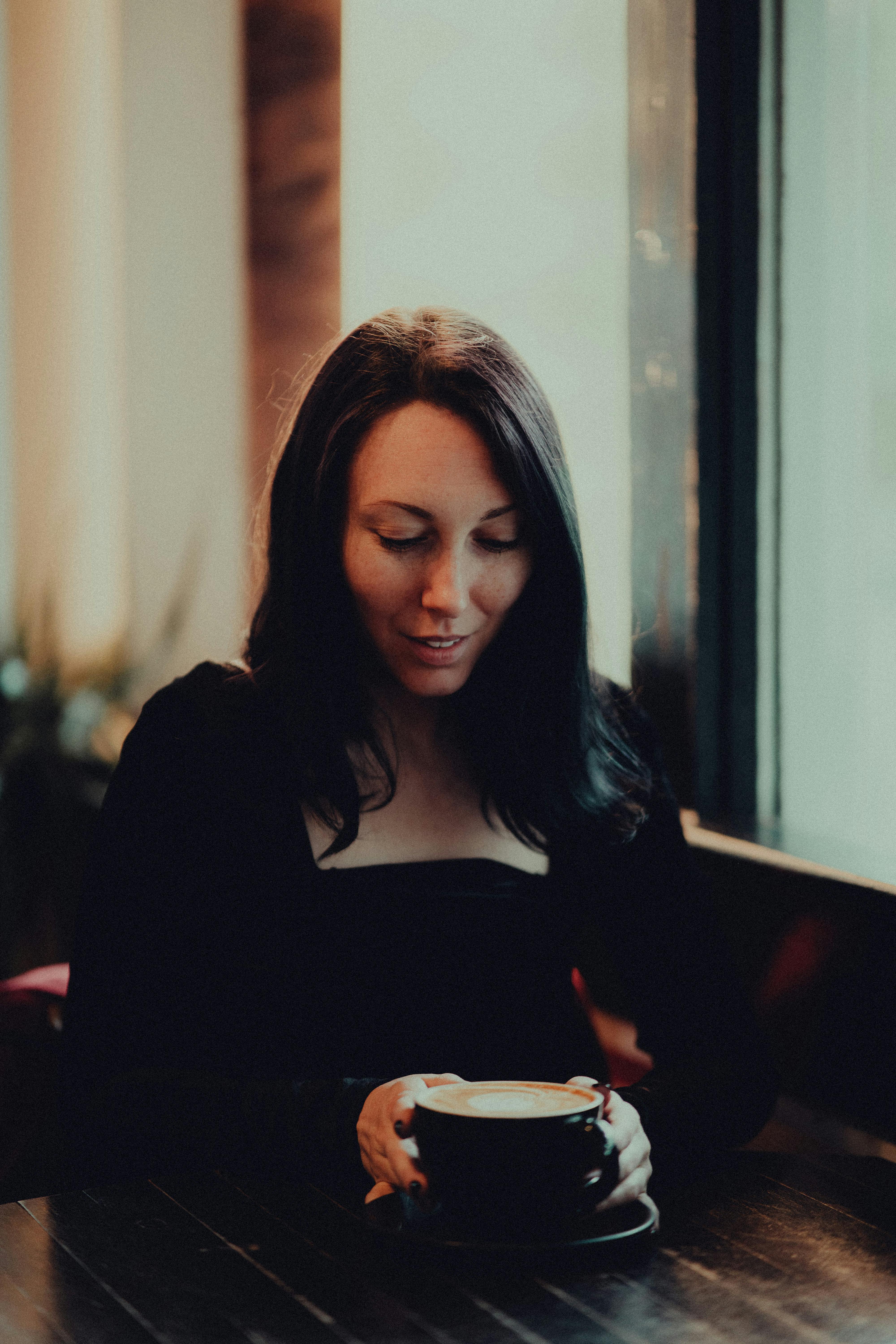 A woman holding a coffee cup while seated indoors by a window in a cozy cafe environment.
