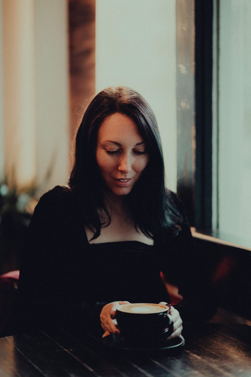 A woman holding a coffee cup while seated indoors by a window in a cozy cafe environment.