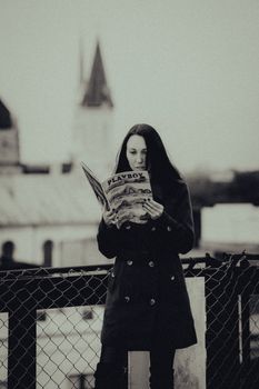 A woman standing outdoors reading a magazine, captured in a monochromatic style with a vintage feel.