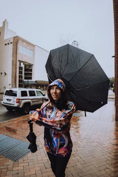 A woman with a colorful hoodie and broken umbrella walks down a rainy city street.