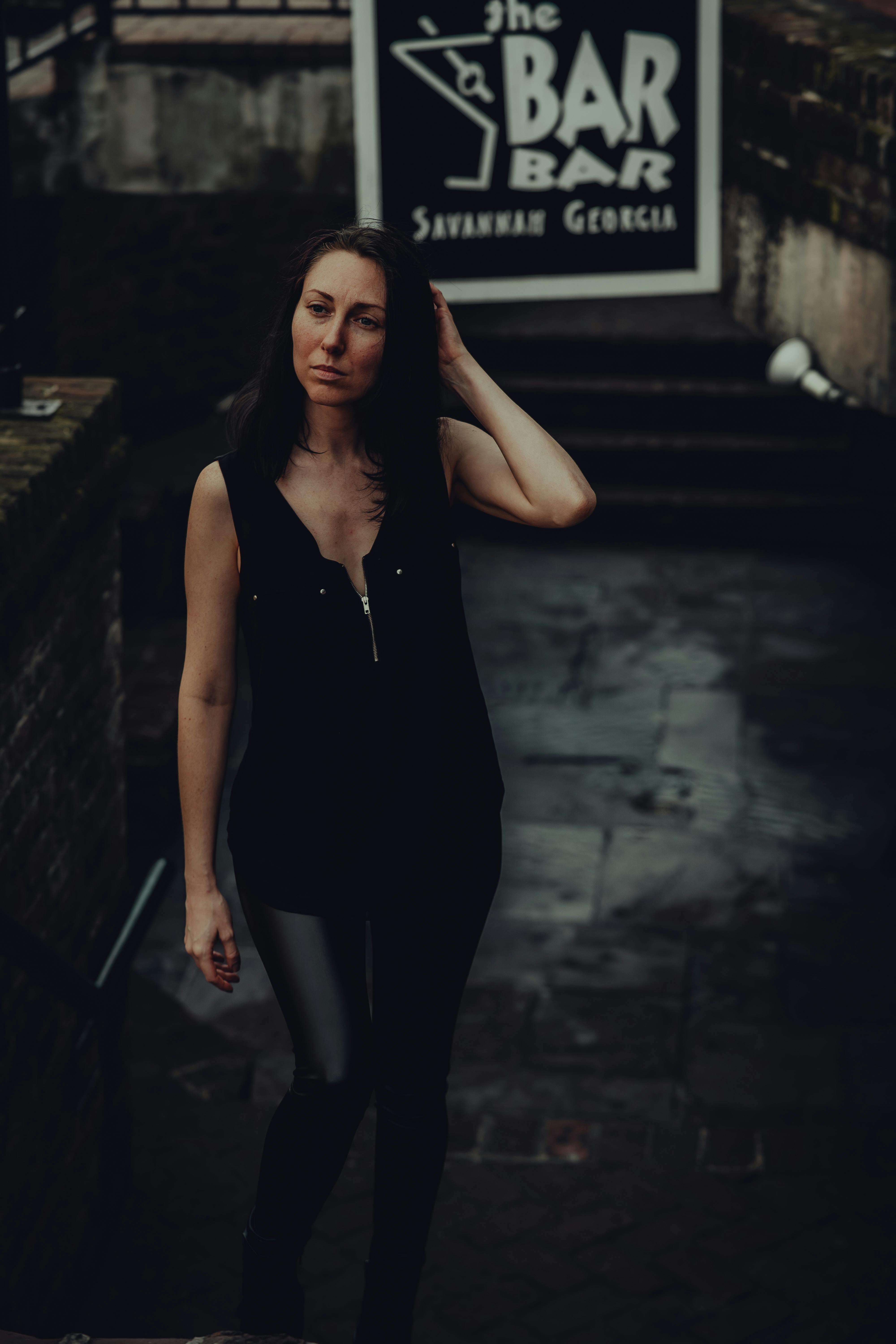 Woman in a black sleeveless top standing by a sign for The Bar Bar in Savannah, Georgia during daytime.