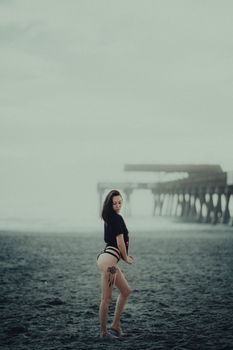 A woman posing on a foggy beach near a wooden pier, wearing a t-shirt and bikini bottoms.