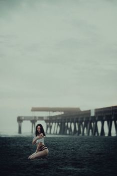 A woman in swimwear poses artistically on a misty beach next to a wooden pier.