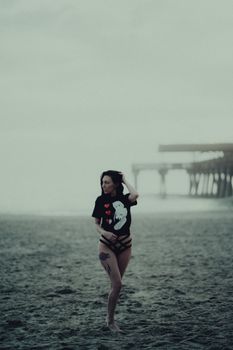 Brunette woman posing on a foggy beach near a pier, wearing a graphic t-shirt and panties.