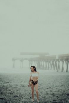 A woman poses barefoot in a tube top on a foggy beach near a wooden pier, creating a moody coastal scene.