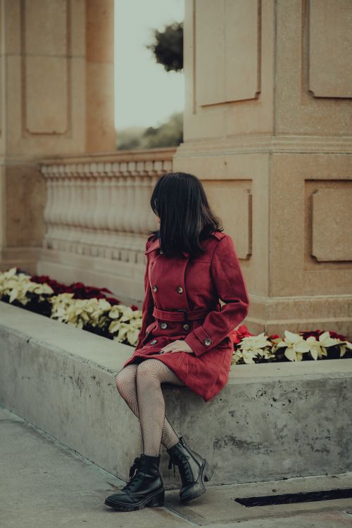 Free A woman in a stylish red coat sitting on a ledge in an outdoor garden setting. Stock Photo