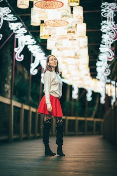 Elegant woman in a red dress posing under colorful lanterns on a wooden path at night.