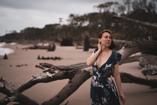 Woman in a floral dress stands on a wild beach with driftwood, exuding natural beauty and serenity.