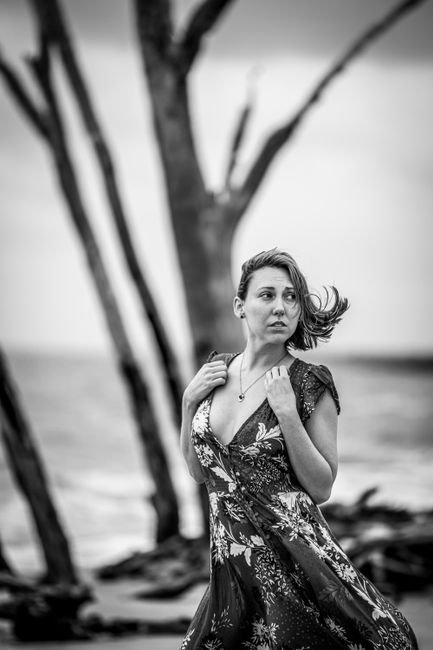 Black and white portrait of a woman in a floral dress by a windy shoreline.