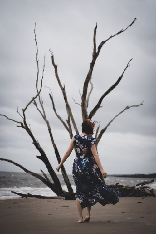 Free A woman in a floral dress strolls along a sandy beach with a bare tree and gray skies. Stock Photo