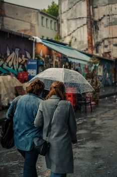 Two women walking under an umbrella in a rainy urban street with graffiti walls.