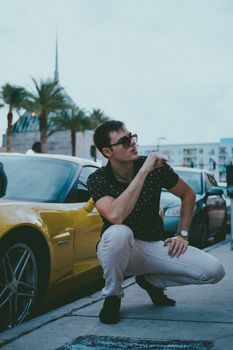 Stylish man crouches by a yellow luxury car on an urban street, exuding modern elegance.