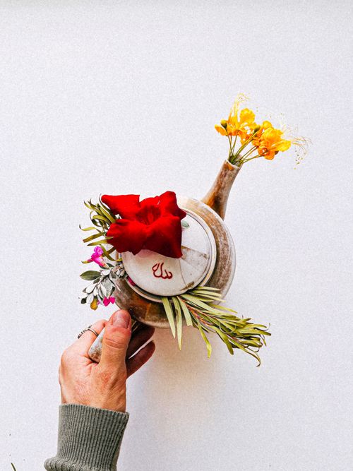 Free Unique floral arrangement in a teapot held by a hand, featuring vibrant red and yellow flowers. Stock Photo