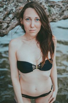 A woman wearing a black bikini poses by a beach tree over water.