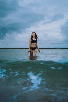 Brunette woman in bikini standing in ocean waves against a cloudy sky.