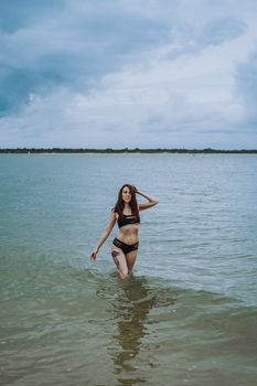 A woman in a black bikini posing confidently in calm ocean waters under a cloudy sky.