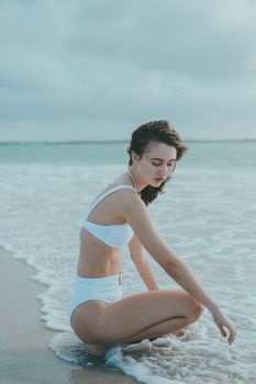 Candid shot of a woman in a white bikini kneeling by the sea shore on a cloudy day.