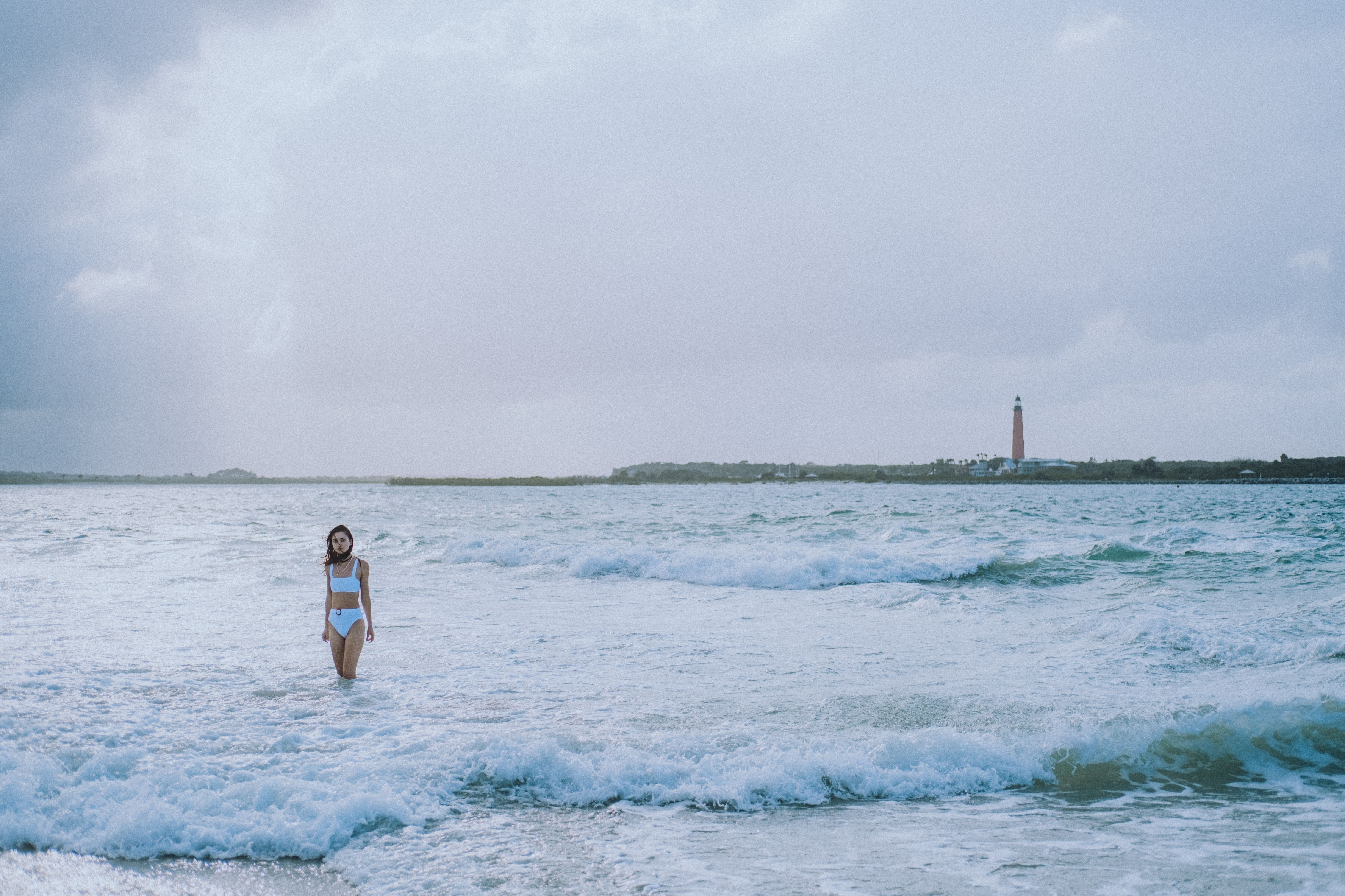 A woman in a white bikini walks along a beach with waves crashing and a lighthouse in the distance.