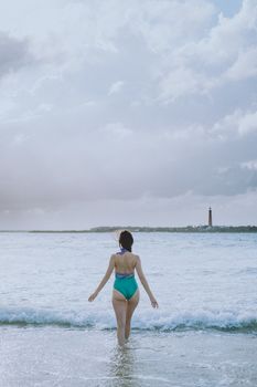 A woman in a swimsuit stands at the shoreline facing the ocean with a distant lighthouse.