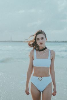 Fashionable young woman in white bikini enjoying a serene beach setting with the sea as backdrop.