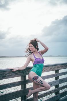 A woman in a colorful swimsuit poses on a wooden railing by the sea under a cloudy sky.