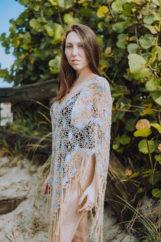 Woman posing in a crochet cover-up on a sandy beach with leafy backdrop.