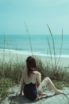 A woman in a swimsuit enjoys a peaceful moment on a sandy beach with grass dunes and gentle waves.
