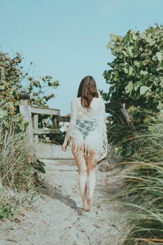 A young woman in a swimsuit and cover-up walks up a sandy path surrounded by greenery.