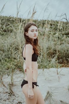 A woman in a black swimsuit stands on a beach with tall grass, gazing into the distance on a cloudy day.